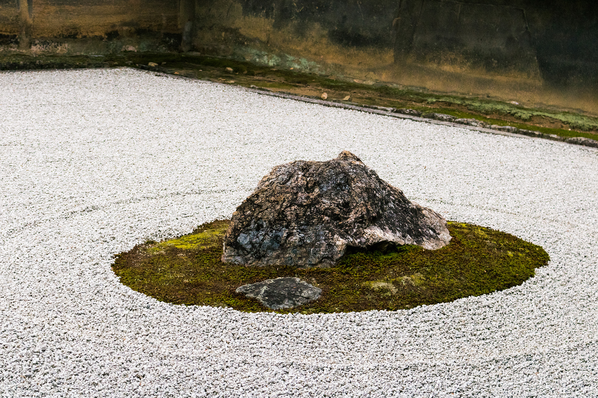 Dark rock surrounded by a small patch of grass, surrounded by small white rocks raked in a zen garden fashion.